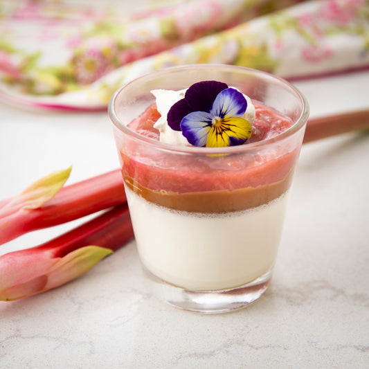 Lovely red Rhubarb branches lay behind a clear cup with a layered dessert: creamy panna cotta, Sunday Night sea salt caramel sauce, rhubarb sauce topped with a purple pansy.