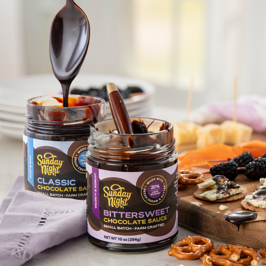 A jar of classic chocolate sauce has a spoon lifting out of it next to a jar of bittersweet chocolate sauce has a spoon resting inside. Both jars sit in front of a cheese tray with pretzels, fruit and drizzled chocolate sauce.