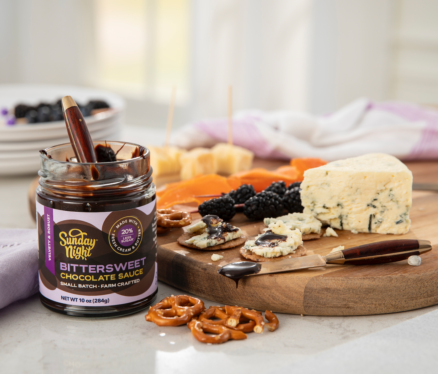 Jar of Sunday Night Bittersweet Chocolate Sauce next to a charcuterie board with cheese, pretzels, and blackberries with chocolate sauce drizzle.